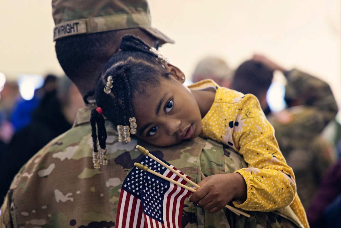 A young girl holding small American flags embraces a soldier as she stares at the camera.