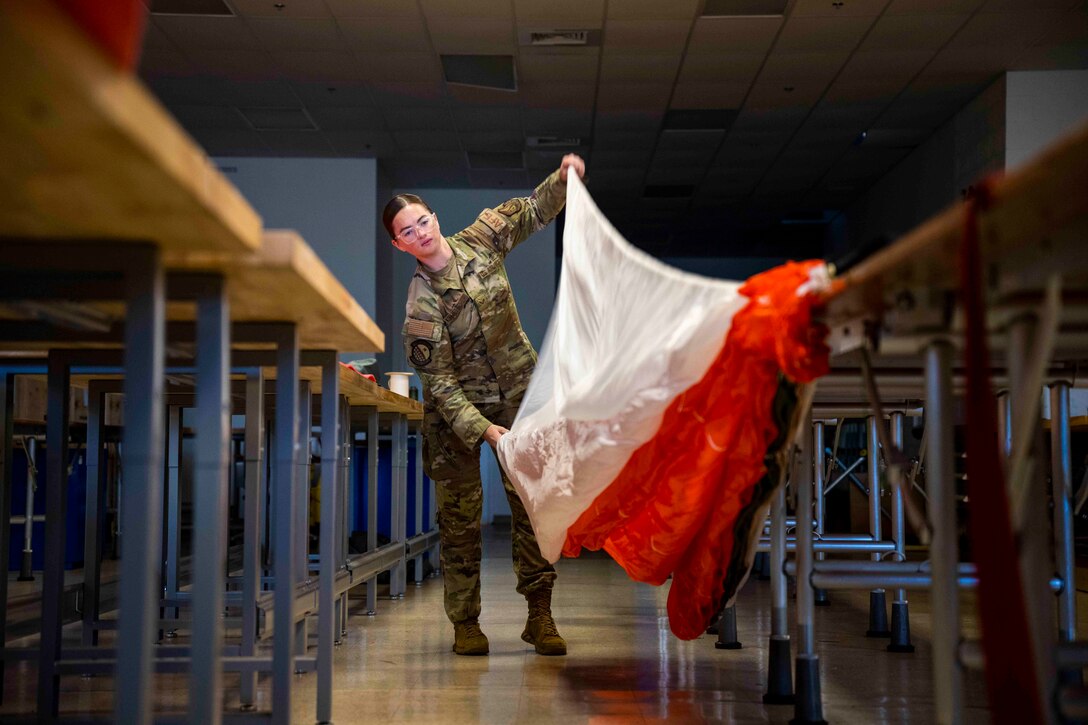 An airman unfurls a parachute while standing between desks.