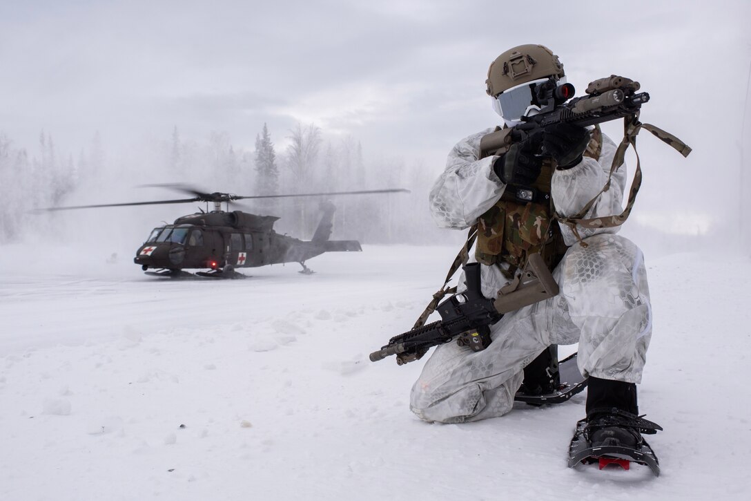 An airman in cold weather gear and snowshoes kneels and aims his weapon while a helicopter lands behind him.