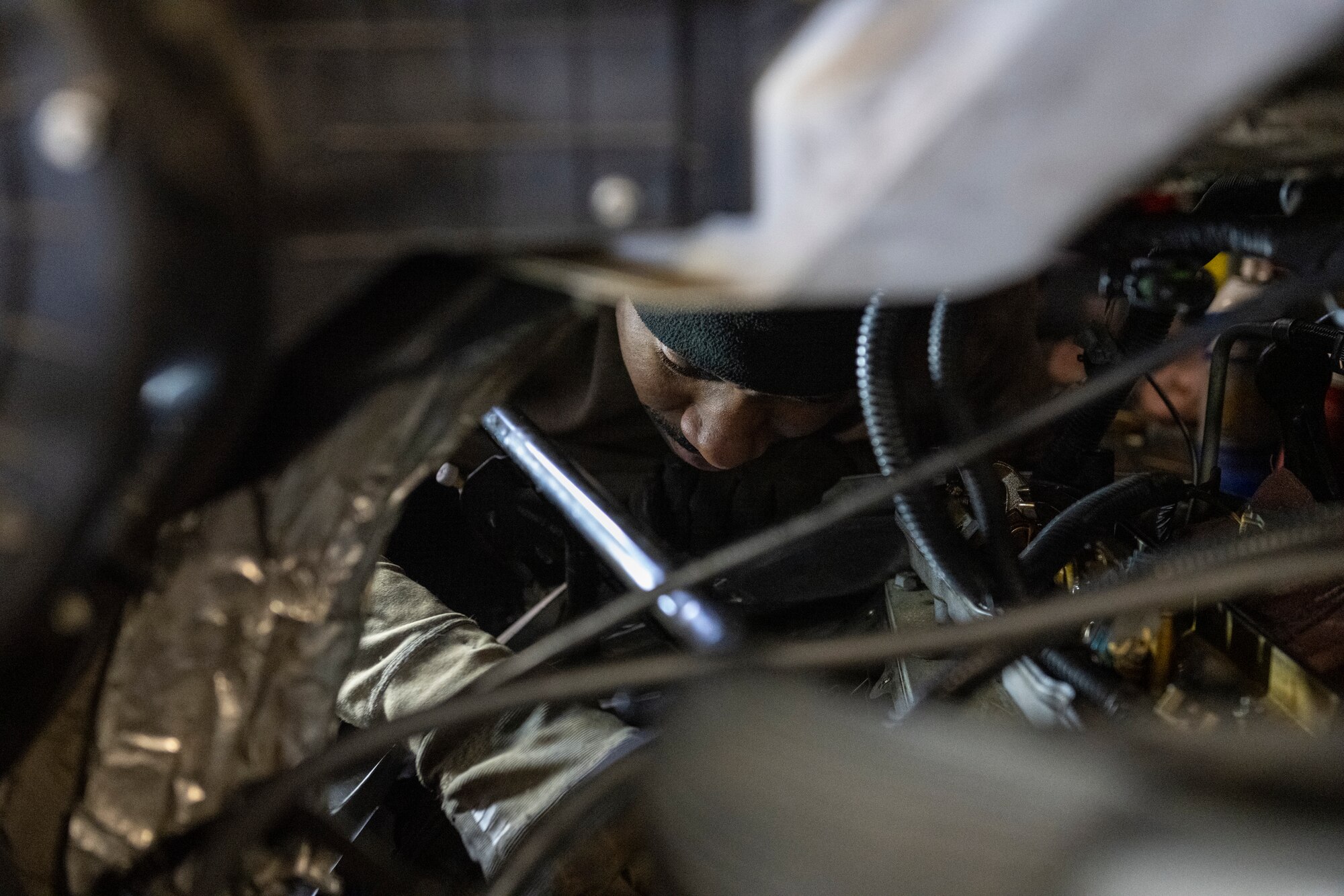 A photo of an Airmen through the wires of a van under the hood.