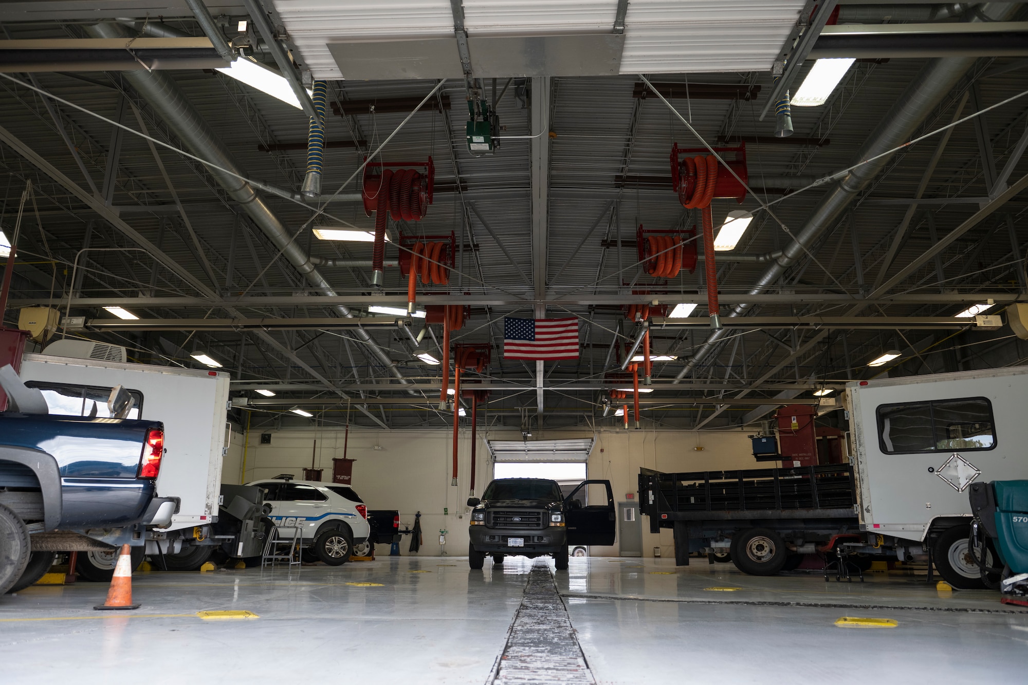 A photo of a car shop with trucks, cop cars, and vans. a truck is parked in the middle and orange pipes hang from the ceiling next to an American flag.