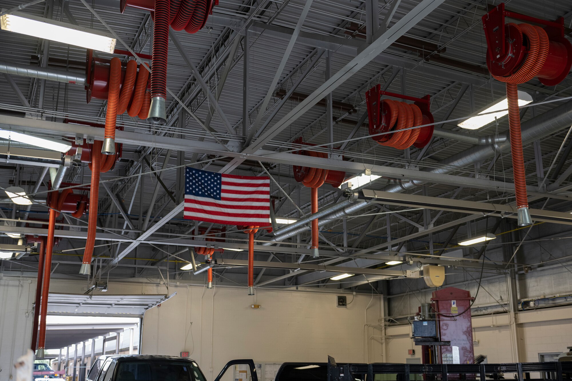 A photo of the ceiling of a big car shop, with an American flag handing in the middle of 10 orange pipes hanging in a row also form the ceiling.