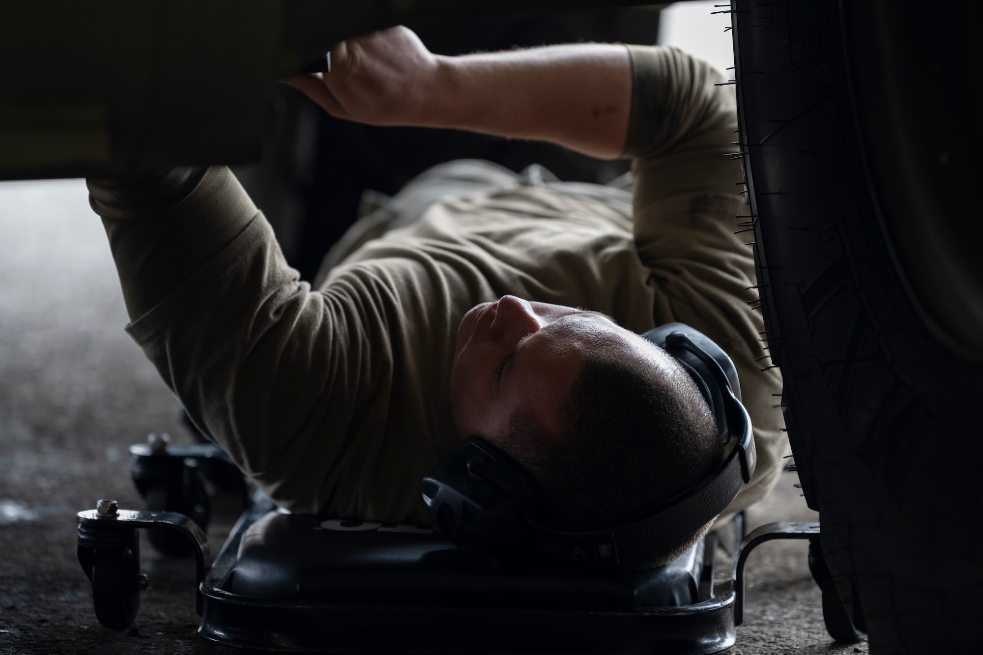 A photo of an Airman laying on his back on a wheeling chair/device reaching into the under carriage of a forklift.