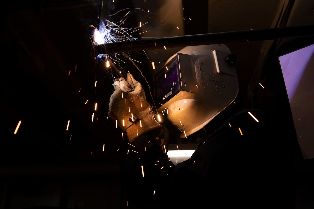 Sparks fly as a welder wearing a helmet works on equipment overhead.