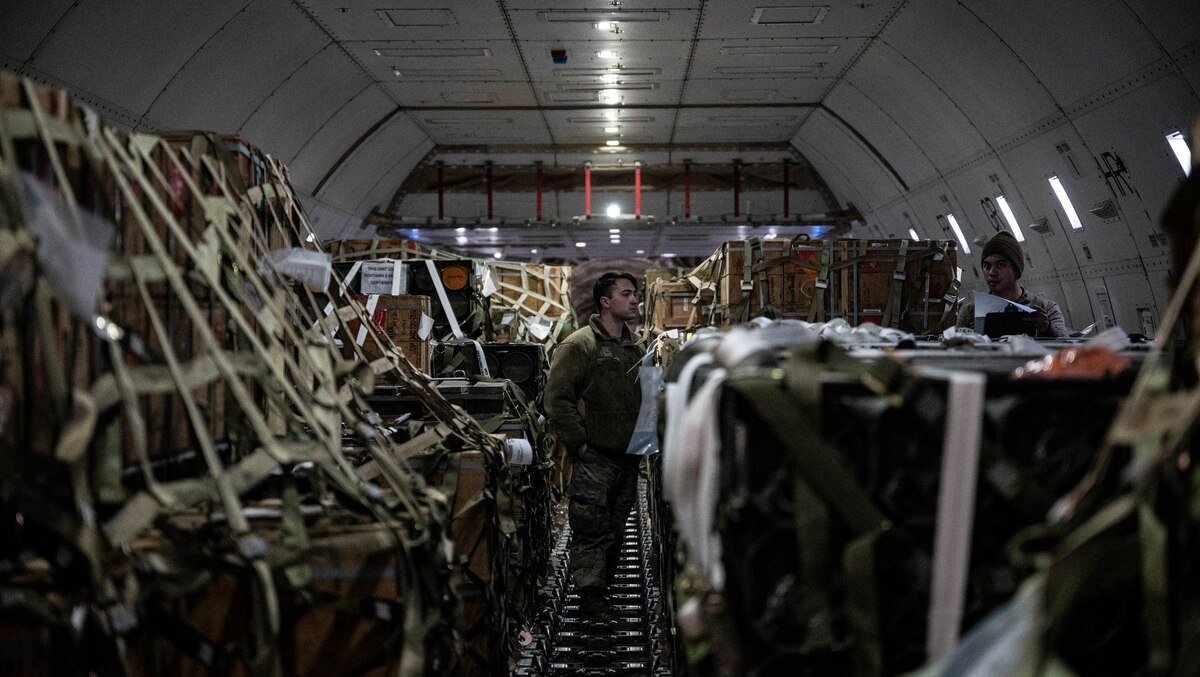 Two men examine cargo inside a small enclosure; one is holding a paper.