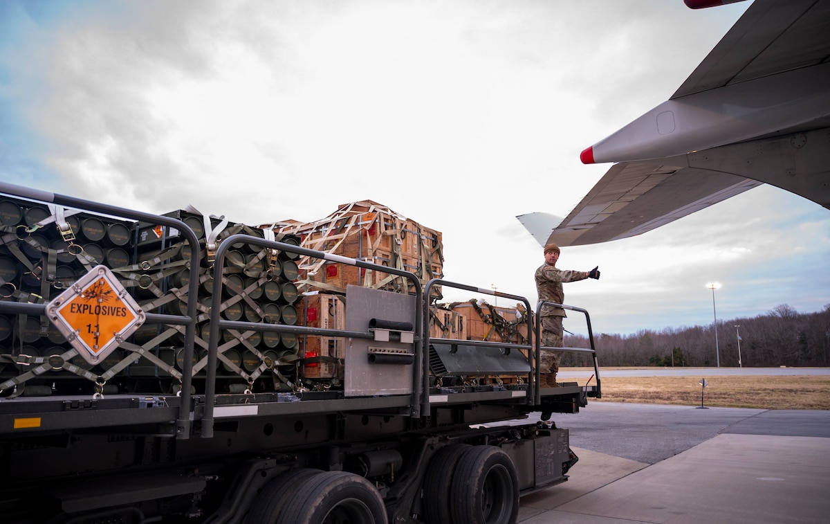 A man standing on the bed of a military vehicle makes a hand gesture to someone who’s off camera. The wing of a plane is in the foreground.