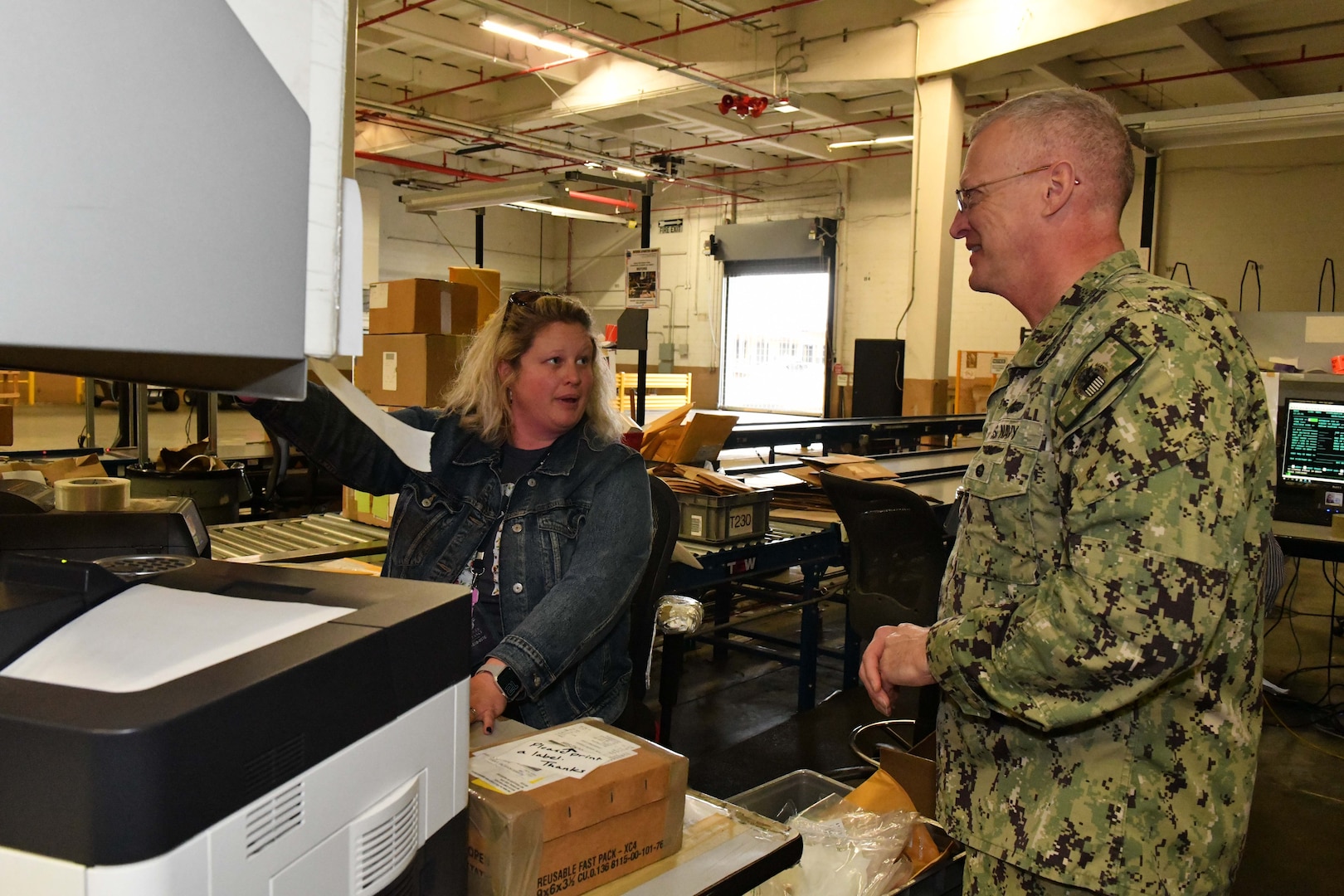 Photo shows man talking to woman sitting at large machine