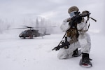 An airman in cold weather gear and snowshoes kneels and aims his weapon while a helicopter lands behind him.