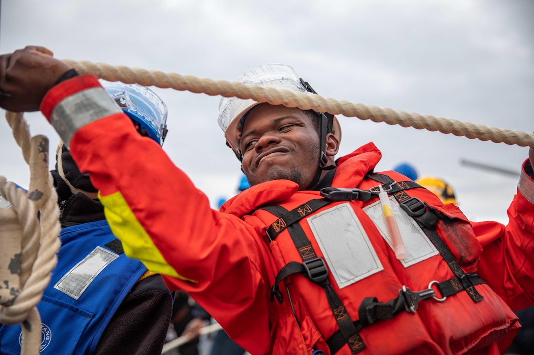 A sailor is shown close-up, pulling a rope.