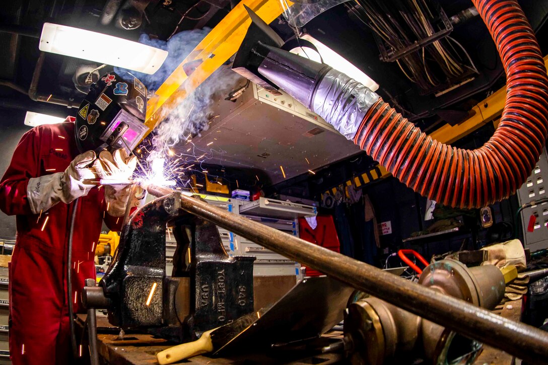 A sailor wearing protective gear welds equipment as sparks fly.