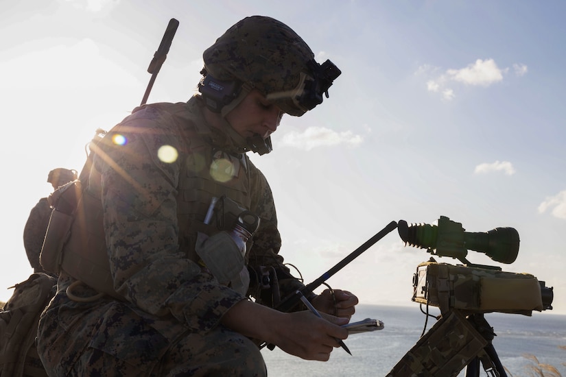 Rays shine over the shoulder of a Marine in a helmet checking data while standing next to target equipment.