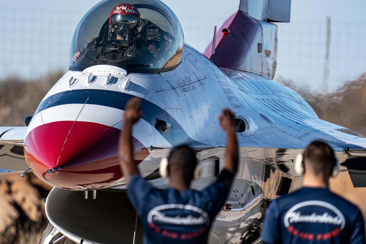 An airman sits in the cockpit of a parked jet as two others look toward them.