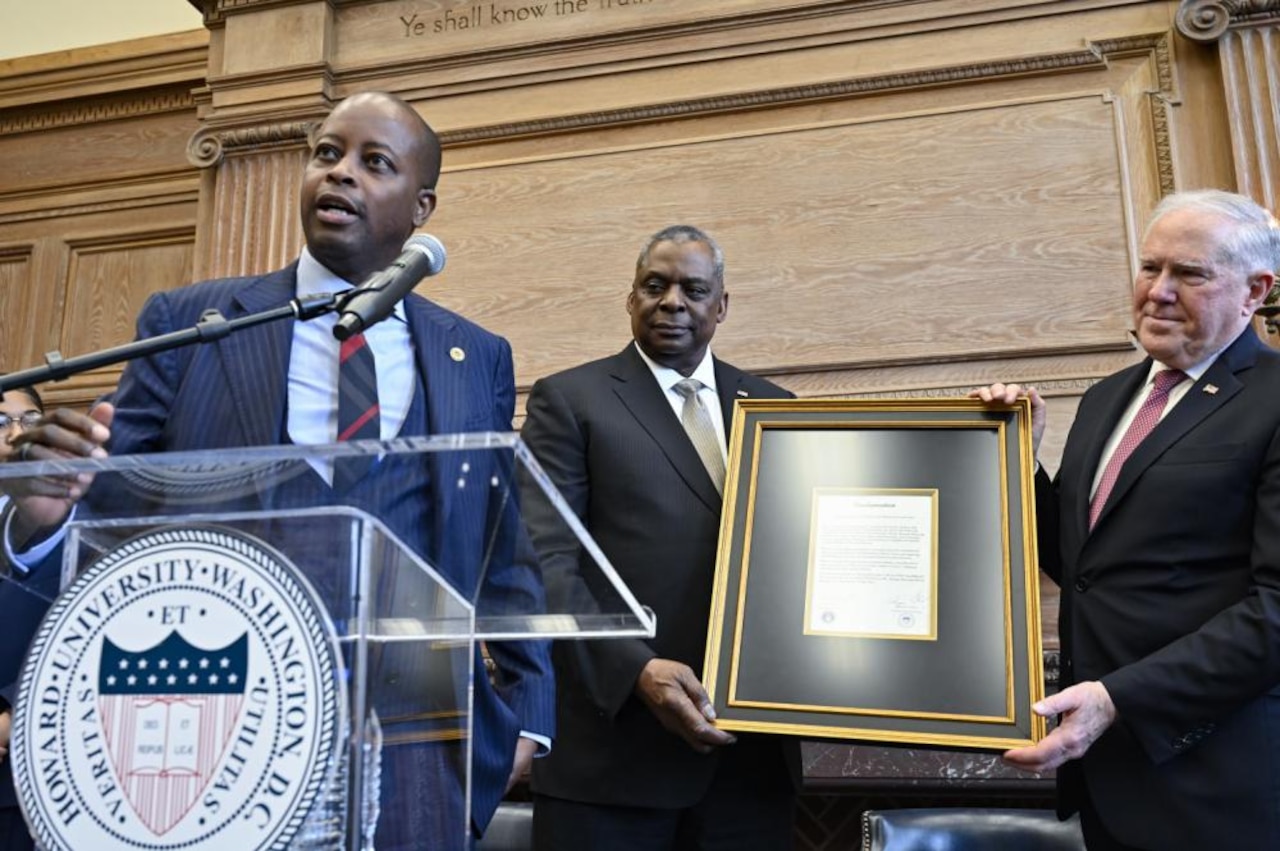 A man stands at a lectern and behind him two men hold a framed proclamation. The seal on the front of the lectern indicates that they are at Howard University.