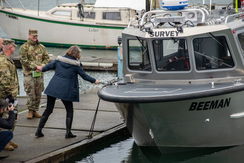 A woman in a dark blue coat standing on a concrete dock smashes a ceremonial champagne bottle over the bow/front of a shiny, steel boat on a cloudy, gray day. A few other people stand around her watching and smiling.