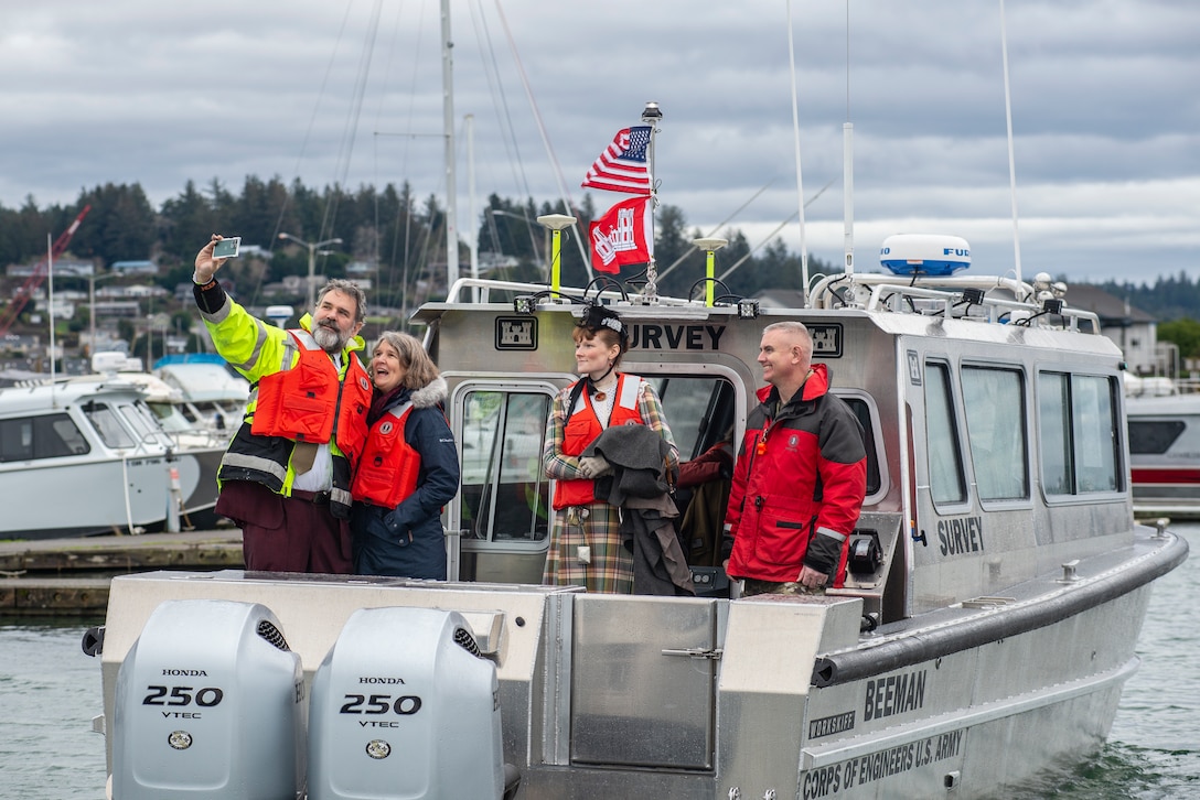 A man and woman take a selfie on the back of a shiny, steel boat on a gray, cloudy day. Another man and woman stand off to the side. Everyone is smiling.