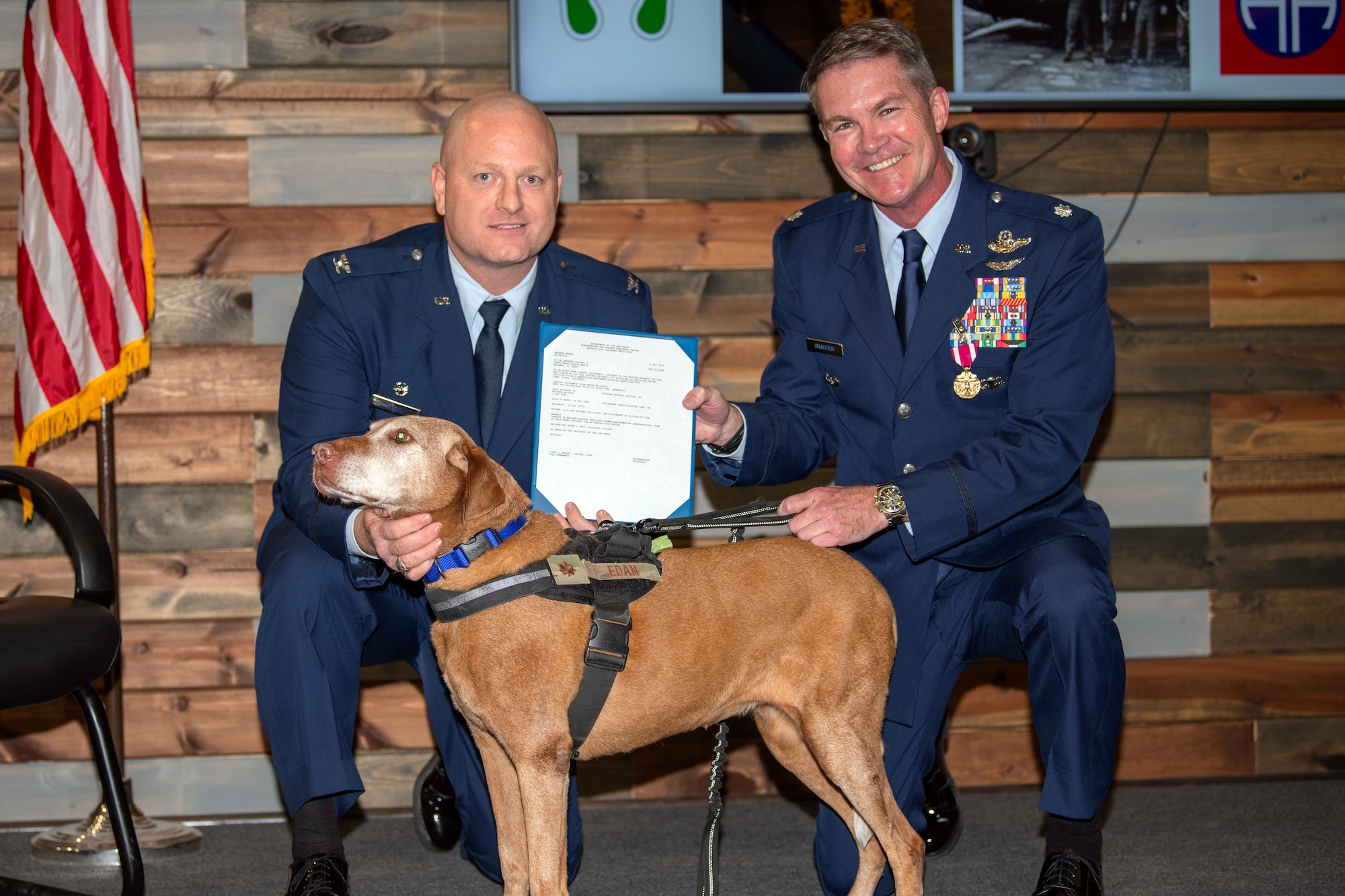 Two Airmen in service dress hold a retirement certificate with a dog in-between them on a wooden stage.