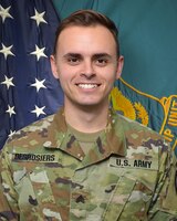 Man wearing U.S. Army uniform standing in front of two flags.