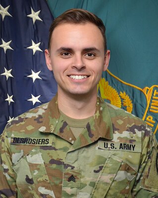 Man wearing U.S. Army uniform standing in front of two flags.