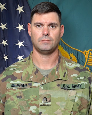 Man in U.S. Army uniform standing in front of two flags.