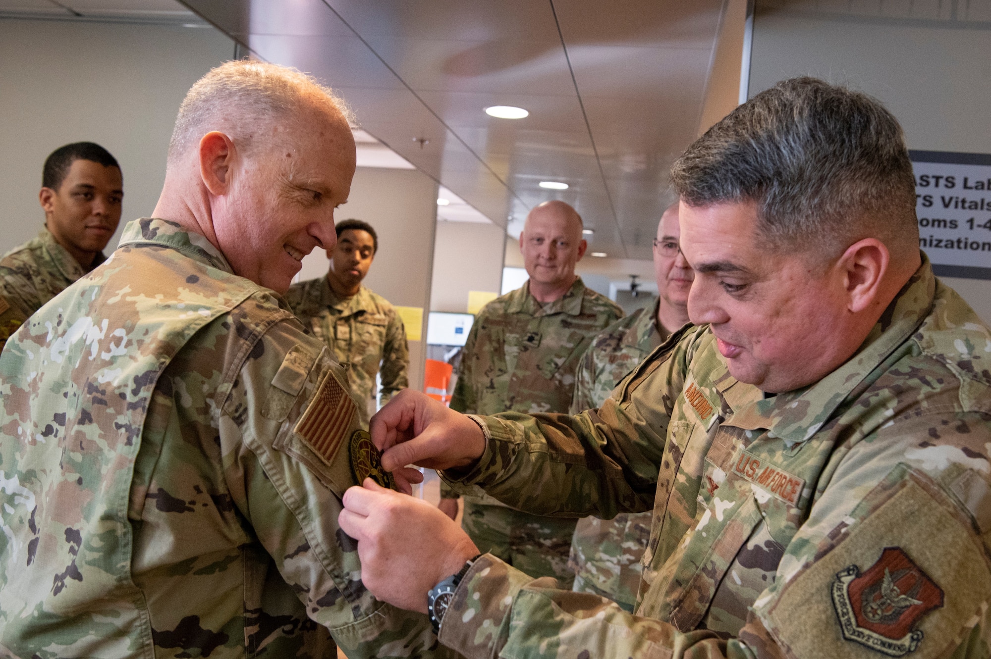 Two men in OCP uniforms interact while one places a patch on the sleeve of the other as onlookers observe