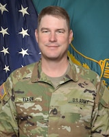 Man in U.S. Army uniform standing in front of two flags.