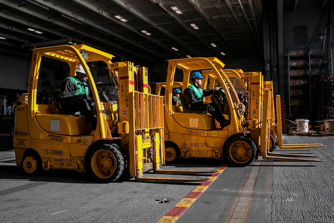 Sailors sit in parked yellow forklifts aboard a ship.