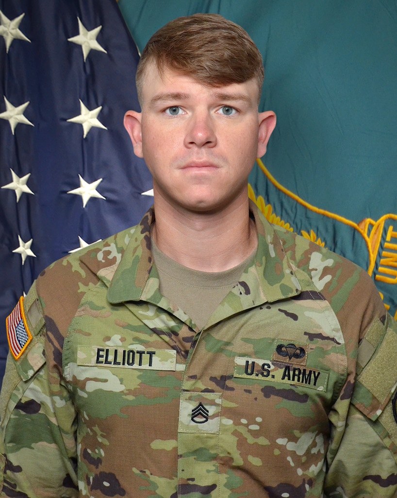 Man in U.S. Army uniform standing in front of two flags.