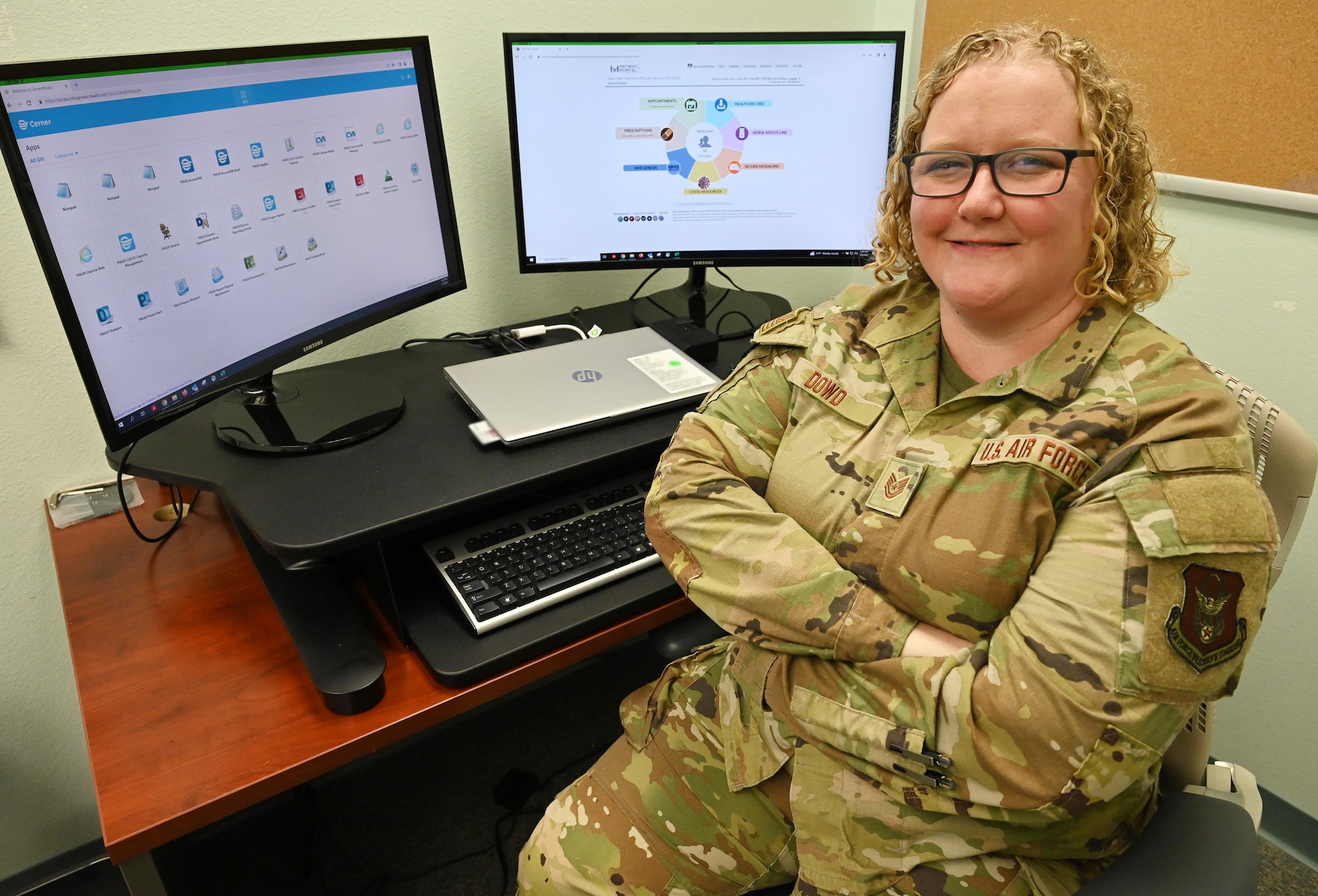 An Airman with arms crossed and seated in an office chair smiles at the viewer, computer monitors behind her display the new electronic health system she implemented.