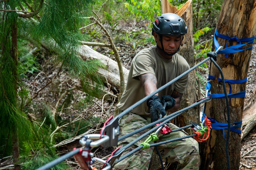 In a wooded area, a soldier wearing a helmet pulls hard on a cord that’s wrapped around a tree.
