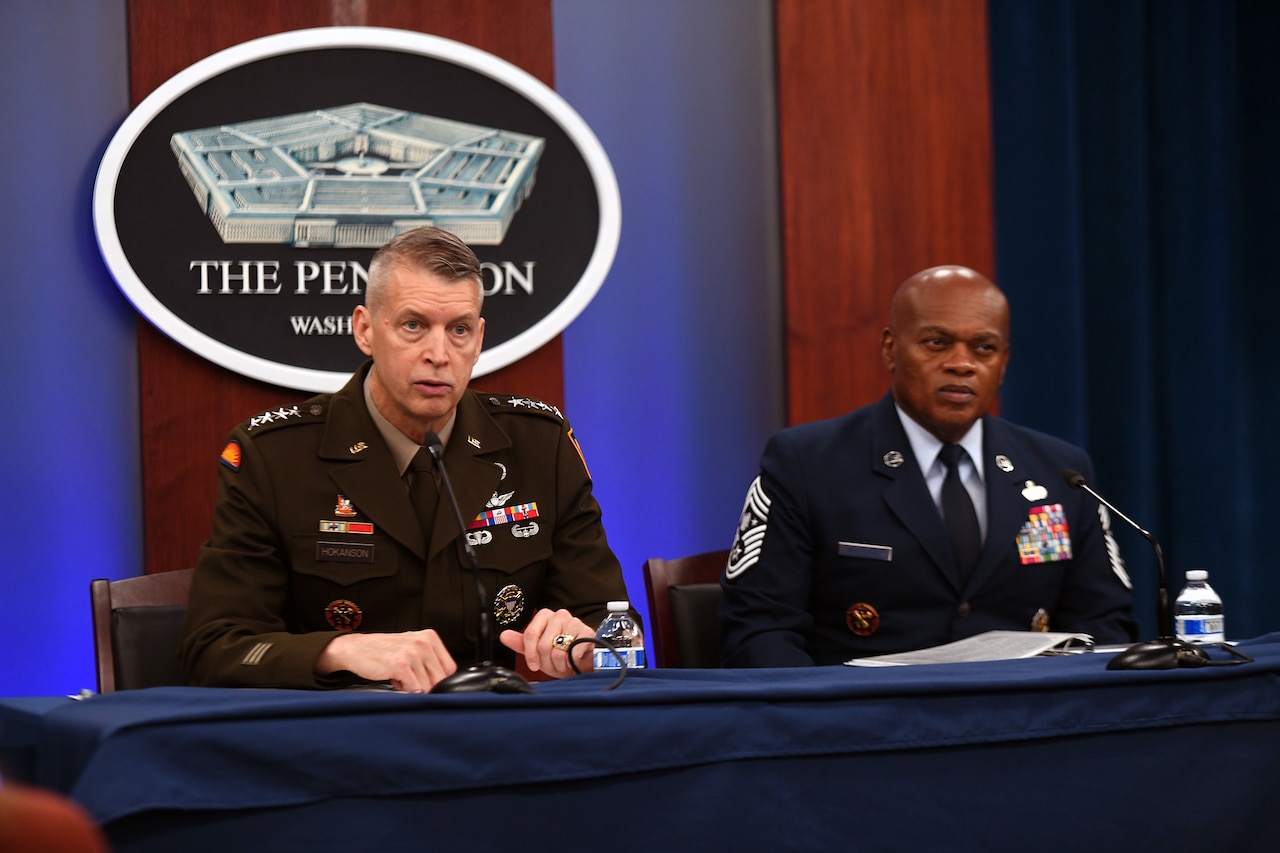 Two men in military uniforms sit behind a table with microphones. The sign behind them indicates that they are at the Pentagon.