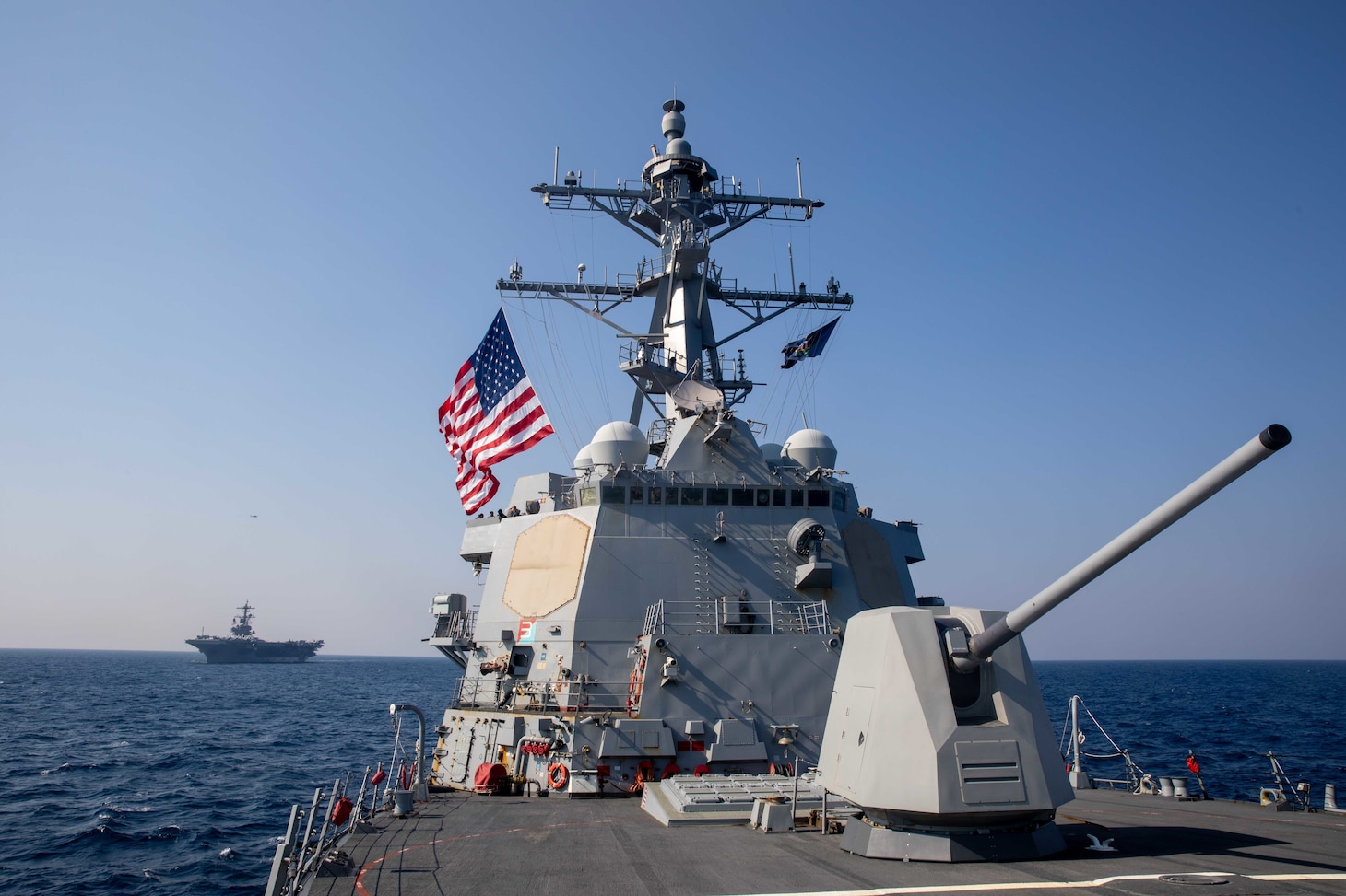 A large Navy vessel sails in the ocean. It is flying a U.S. flag. Another large vessel is far behind it.