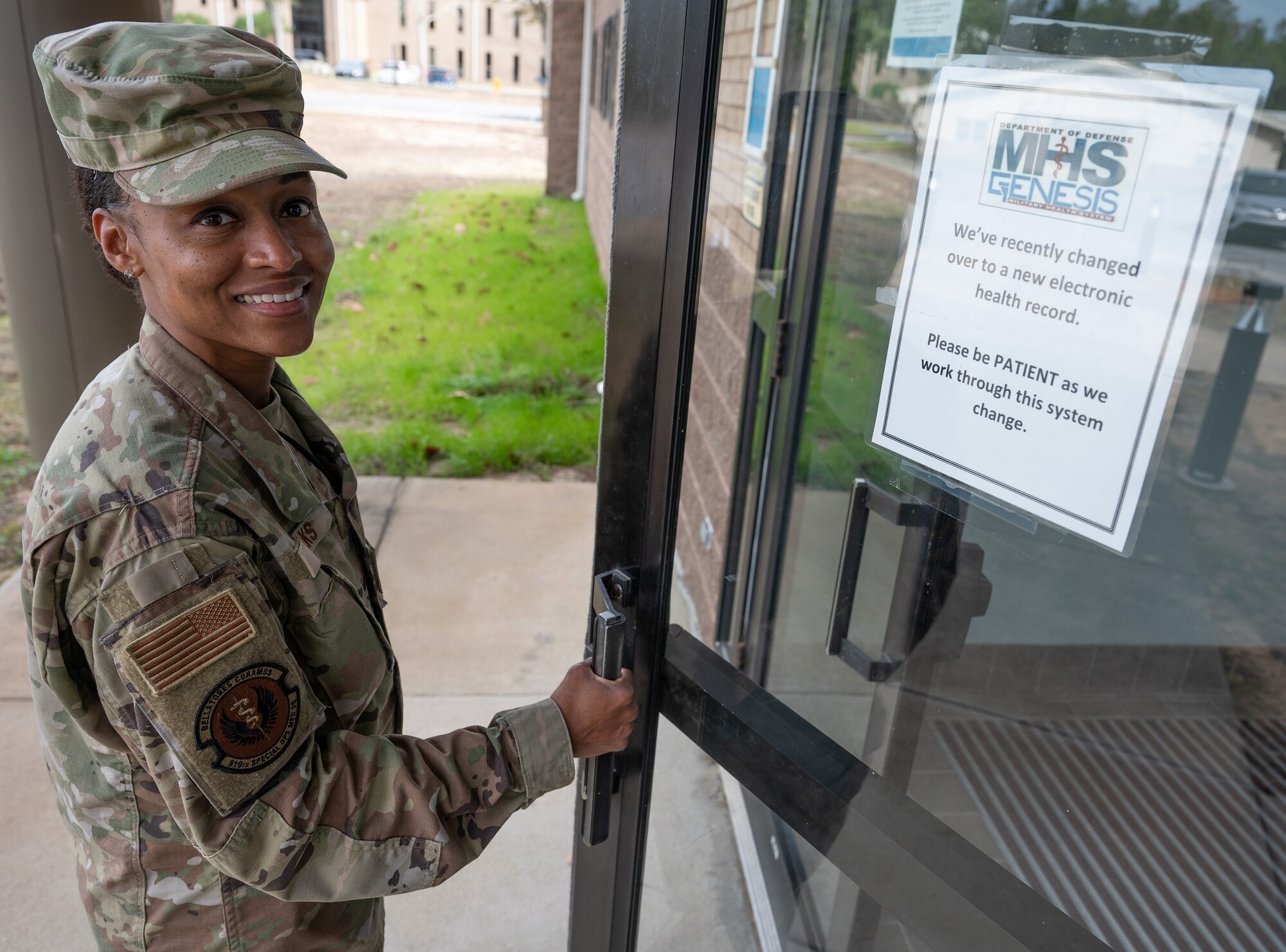 An Airman smiles at the viewer and opens the door of a building with a sign on it announcing the new MHS GENESIS electronic health records system