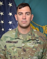 Man in U.S. Army uniform standing in front of two flags.