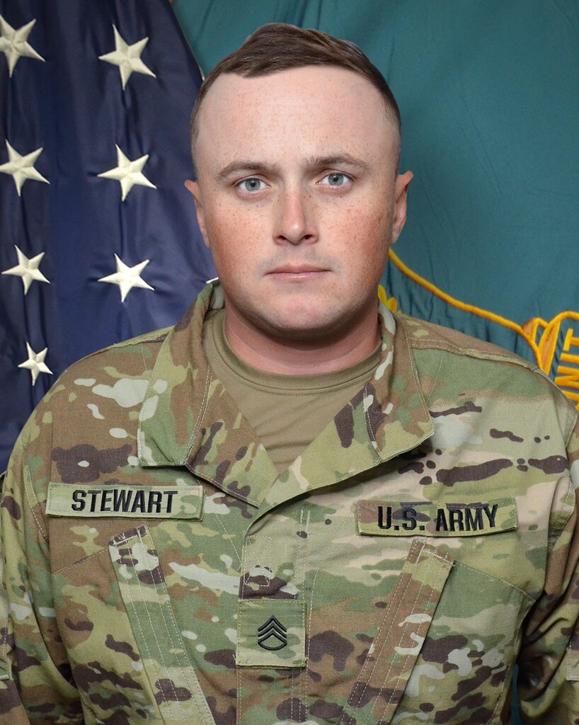Man in U.S. Army uniform standing in front of two flags.