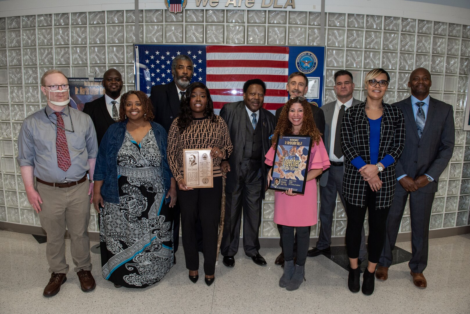 A large group of people posing for a photo. Men and women, dark skinned and light skinned. Two women hold a plaque and a poster.