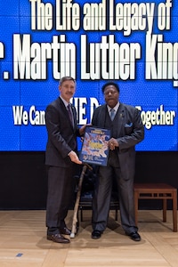 Two men in suits hold a poster on stage. One is light skinned and the other is dark skinned. The dark skinned man has a cane.