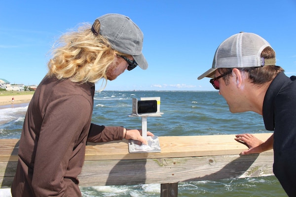 A woman and a man, standing over a peer where a beach and an ocean meet. Between them sits a white cell phone on a mount. The cell phone is recording data reading from the beach.