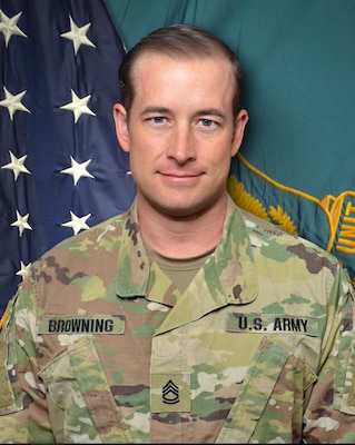 Man in U.S. Army uniform standing in front of two flags.