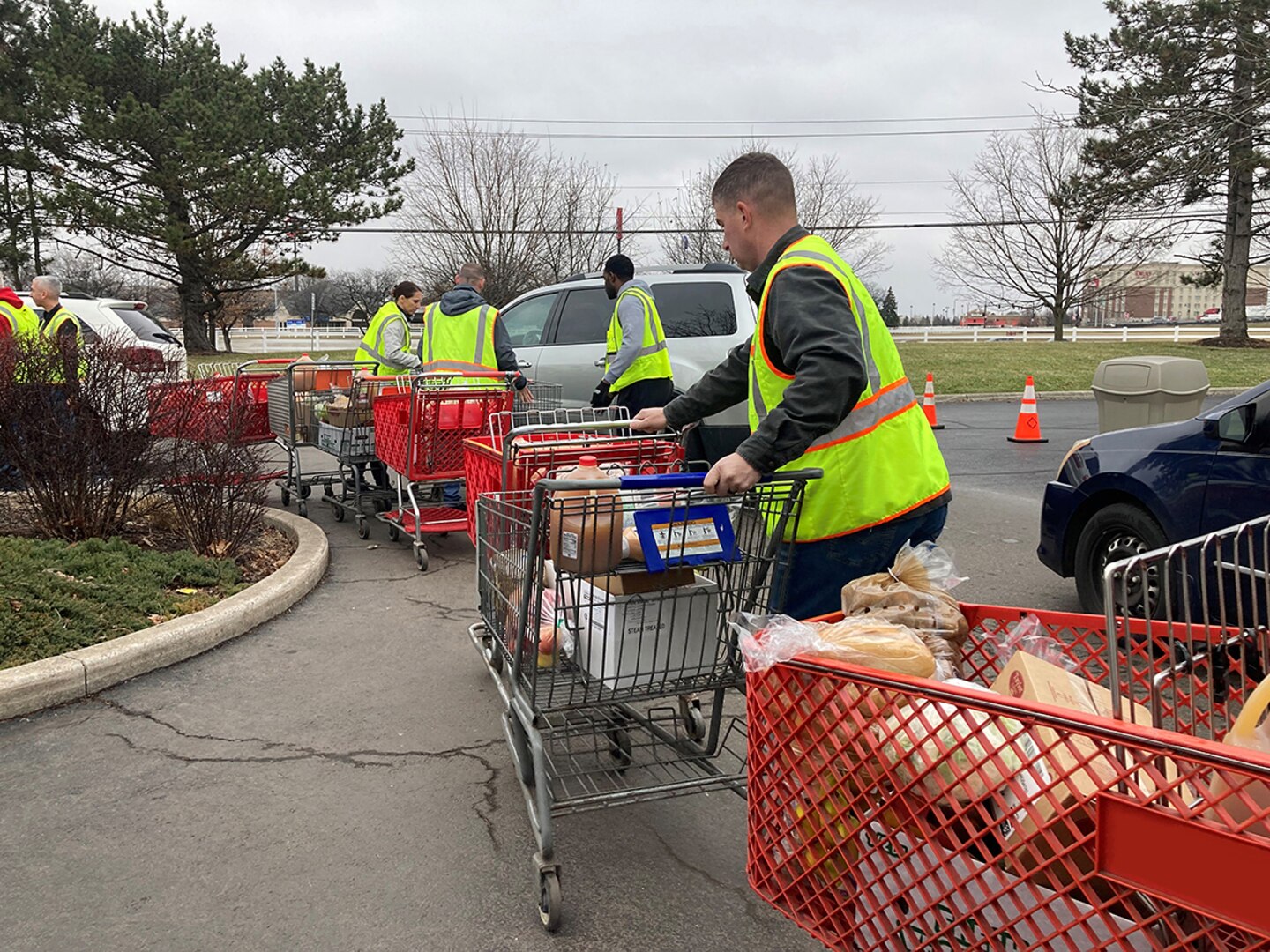 Several men wearing reflective vests handle shopping carts full of food in a outdoor food distribution area in a parking lot.