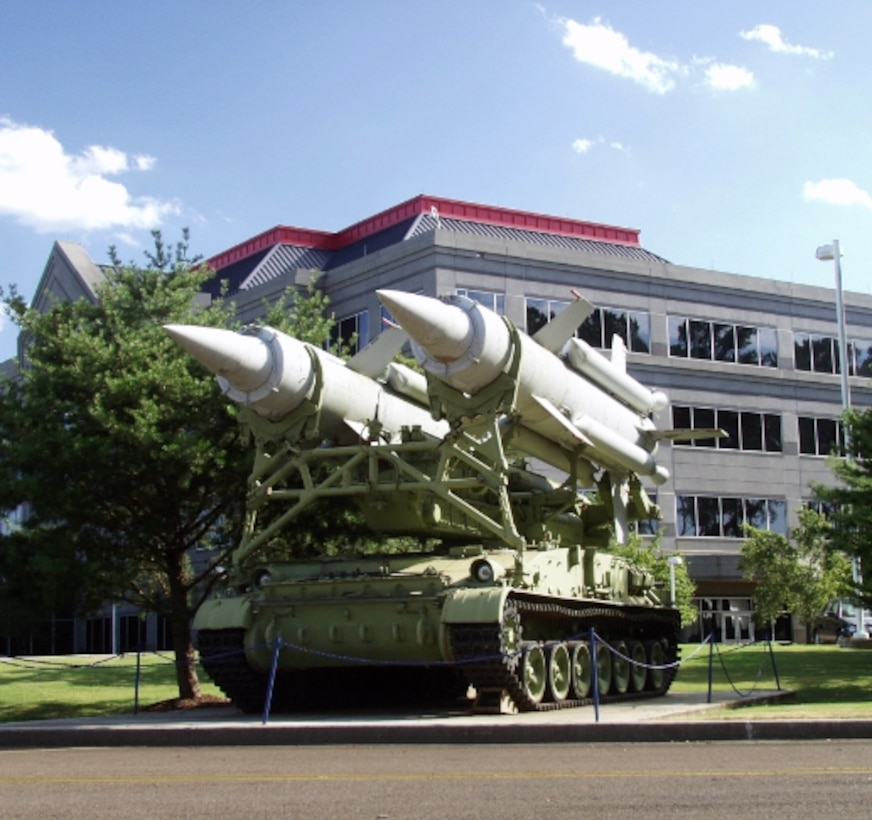 Image of missile-launching vehicle display, outdoors in front of a building, behind a short, chain guard.