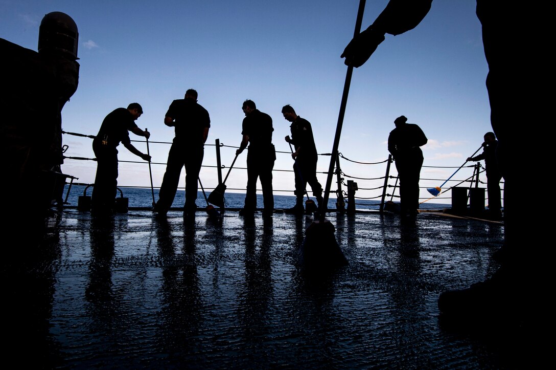 Sailors shown in silhouette against a dark blue sky sweep a ship's deck while at sea.