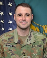 Man in U.S. Army uniform standing in front of two flags.