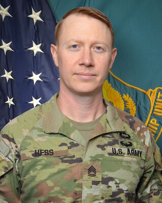 Man in U.S. Army uniform standing in front of two flags.