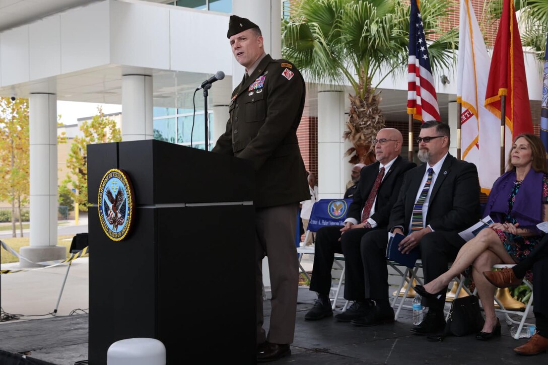 Army brigadier general at podium with US Flag and seated dignitaries behind him.