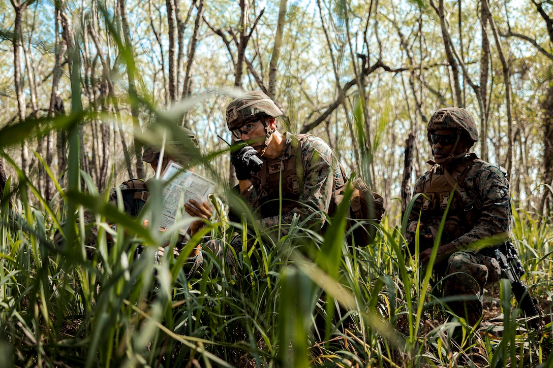 A Marine speaks into a radio while crouched in a forested area with two fellow Marines.