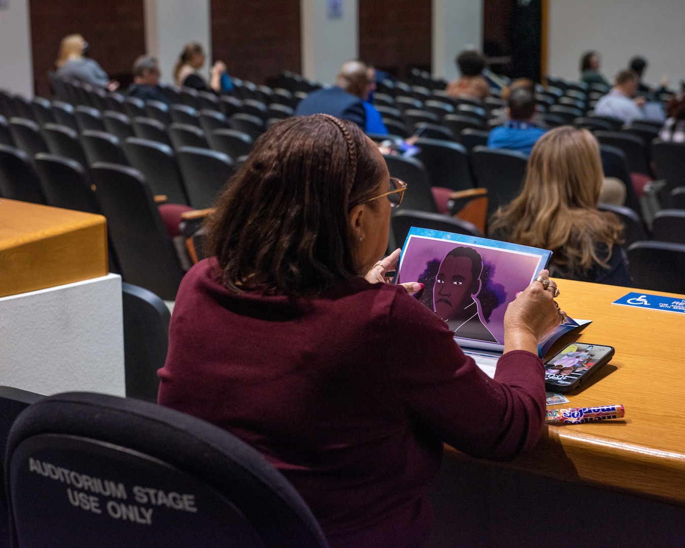 A dark skinned woman wearing a purple sweater looks at a glossy calendar in an auditorium.