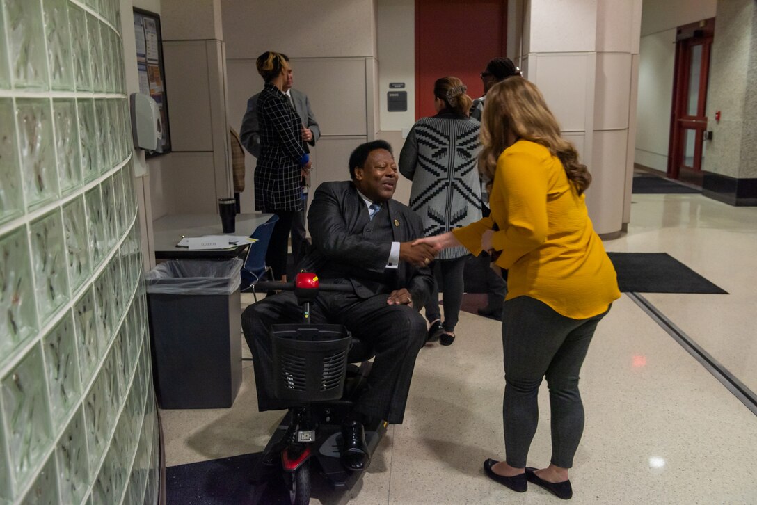 A woman and a man shake hands. One is light skinned and the other dark skinned. The dark skinned man is in a scooter and the other is standing. Many others stand around them.