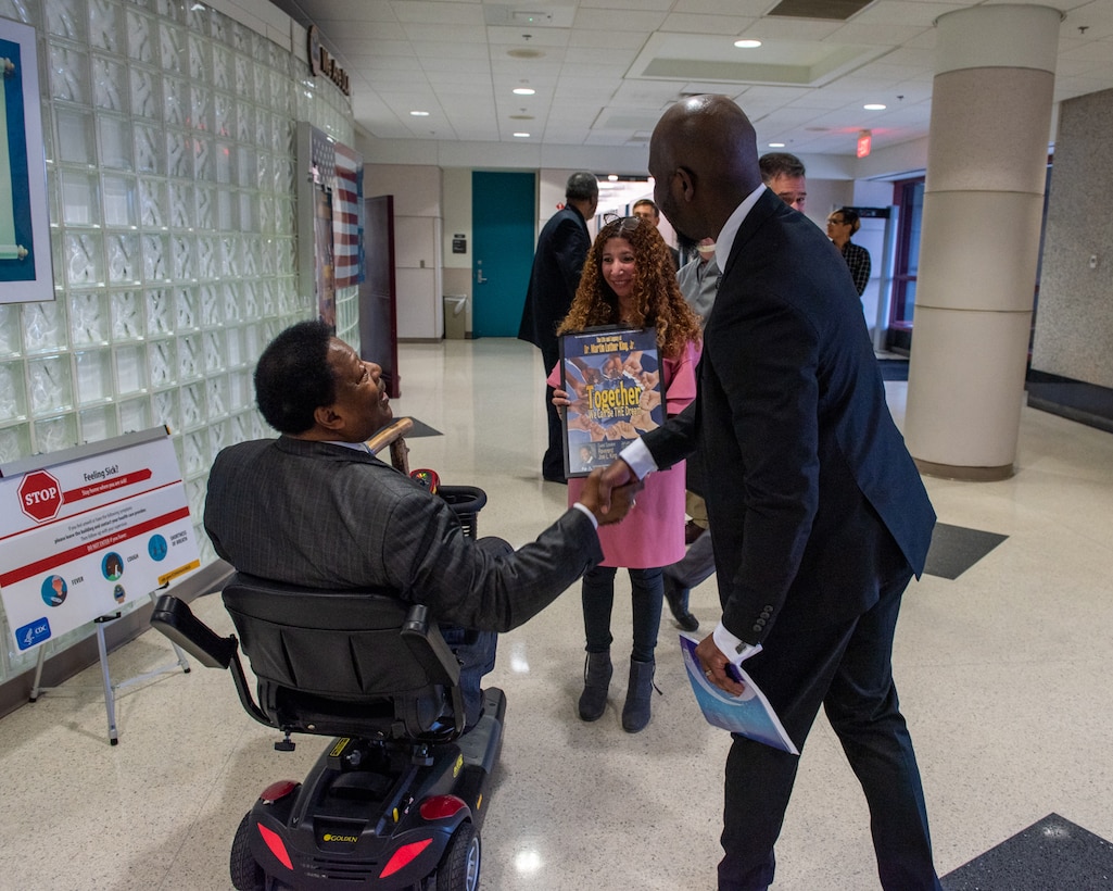Two men shake hands. Both are dark skinned. One is in a scooter and the other is standing. A woman in a pink dress with red hair is in the background holding a framed poster.
