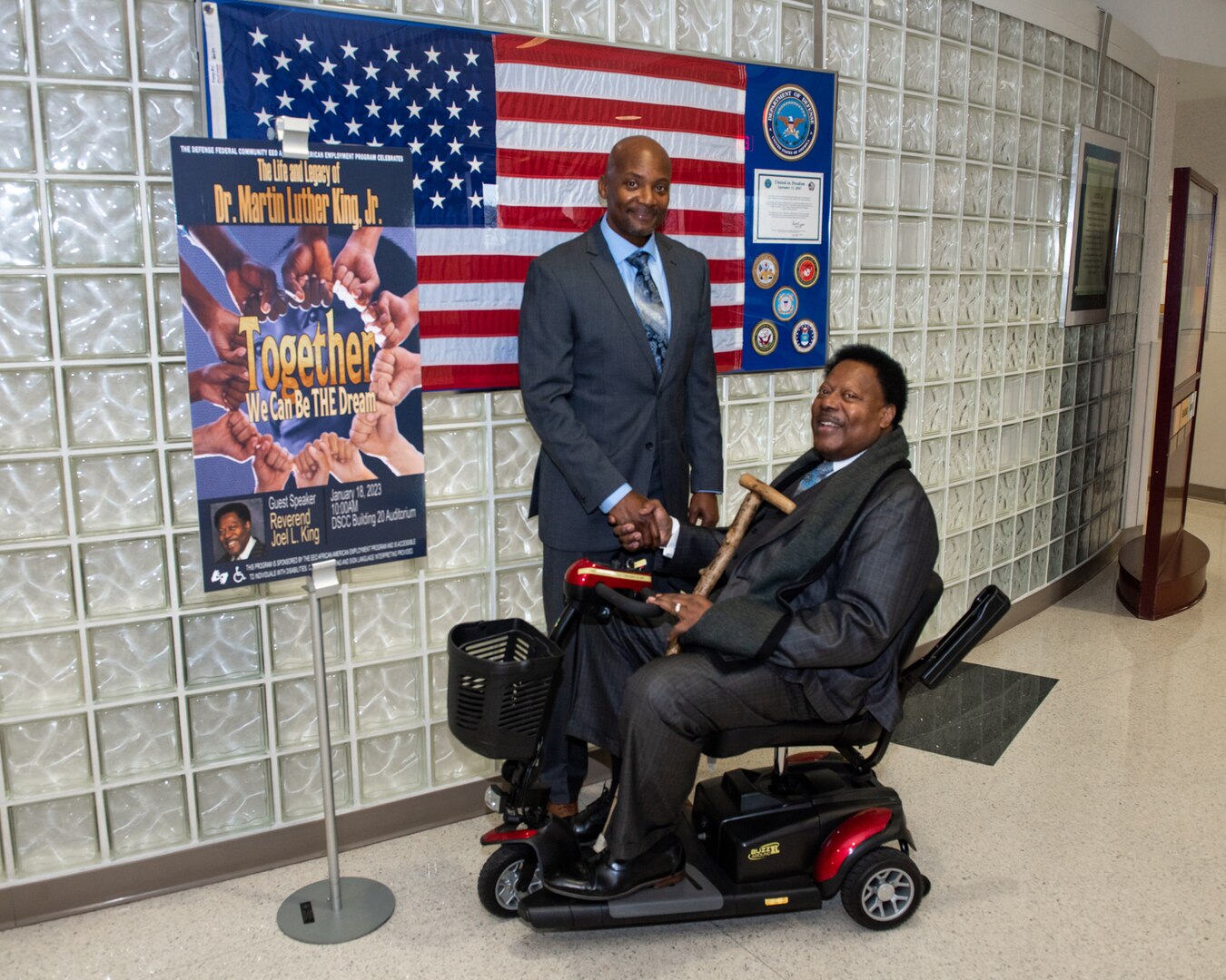 Two dark skinned men shake hands in a hallway. One is seated in a scooter and the other is standing.