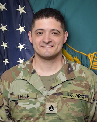 Man in U.S. Army uniform standing in front of two flags.